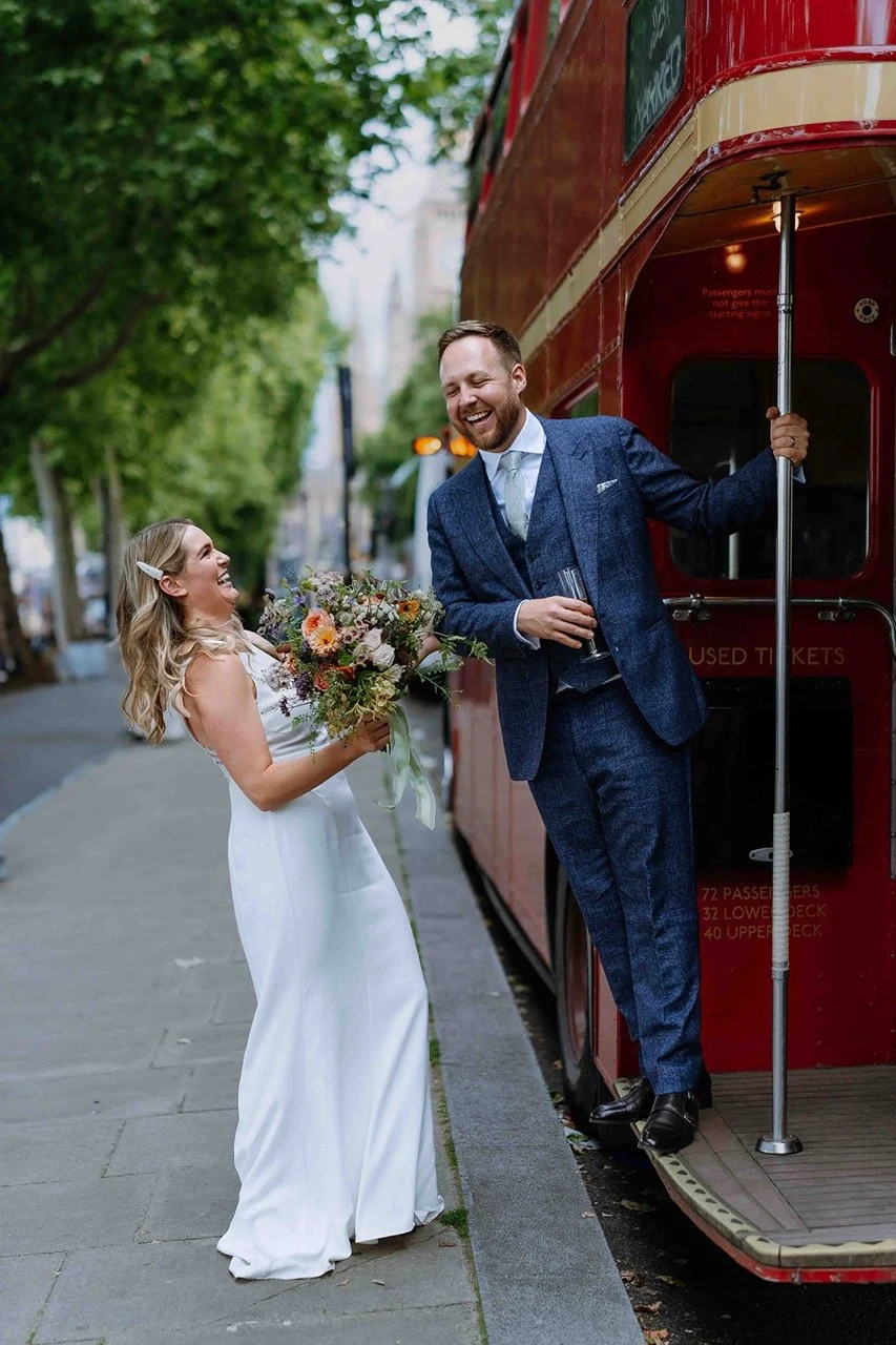 A joyful bride holding a bouquet and a smilling groom standing on a london double-decker bus, with trees and city street in the background.