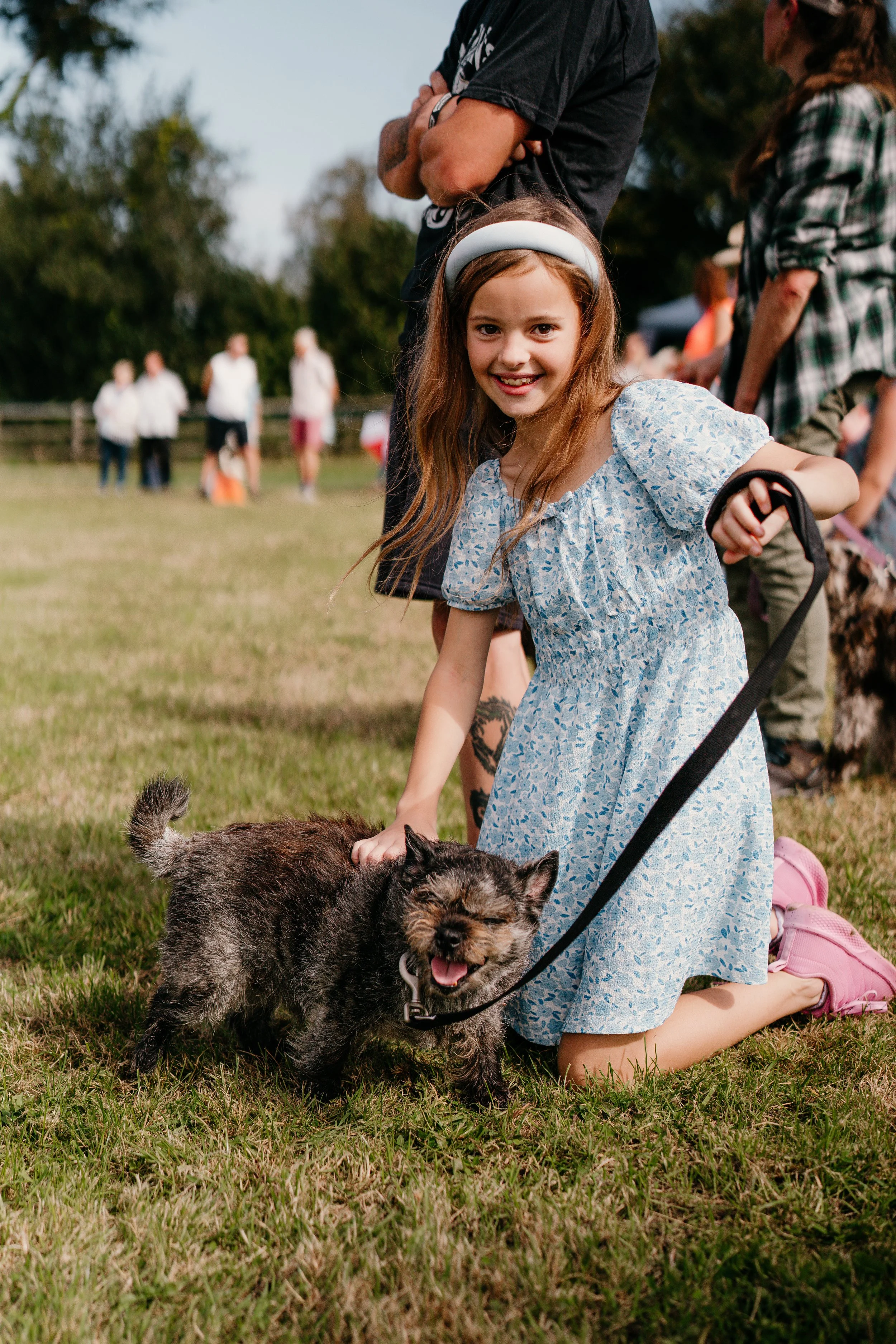A young girl kneeling on the grass, smiling, petting a small scruffy dog with a happy expression, outdoors at a gathering with many people in the background.