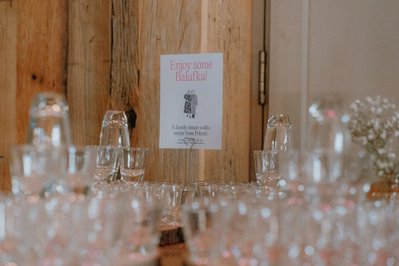 A table set with upside-down glasses and a sign that reads, 'Enjoy some Balufka! A family lemon vodka recipe from Poland.' The sign is on a stand against a wooden wall.