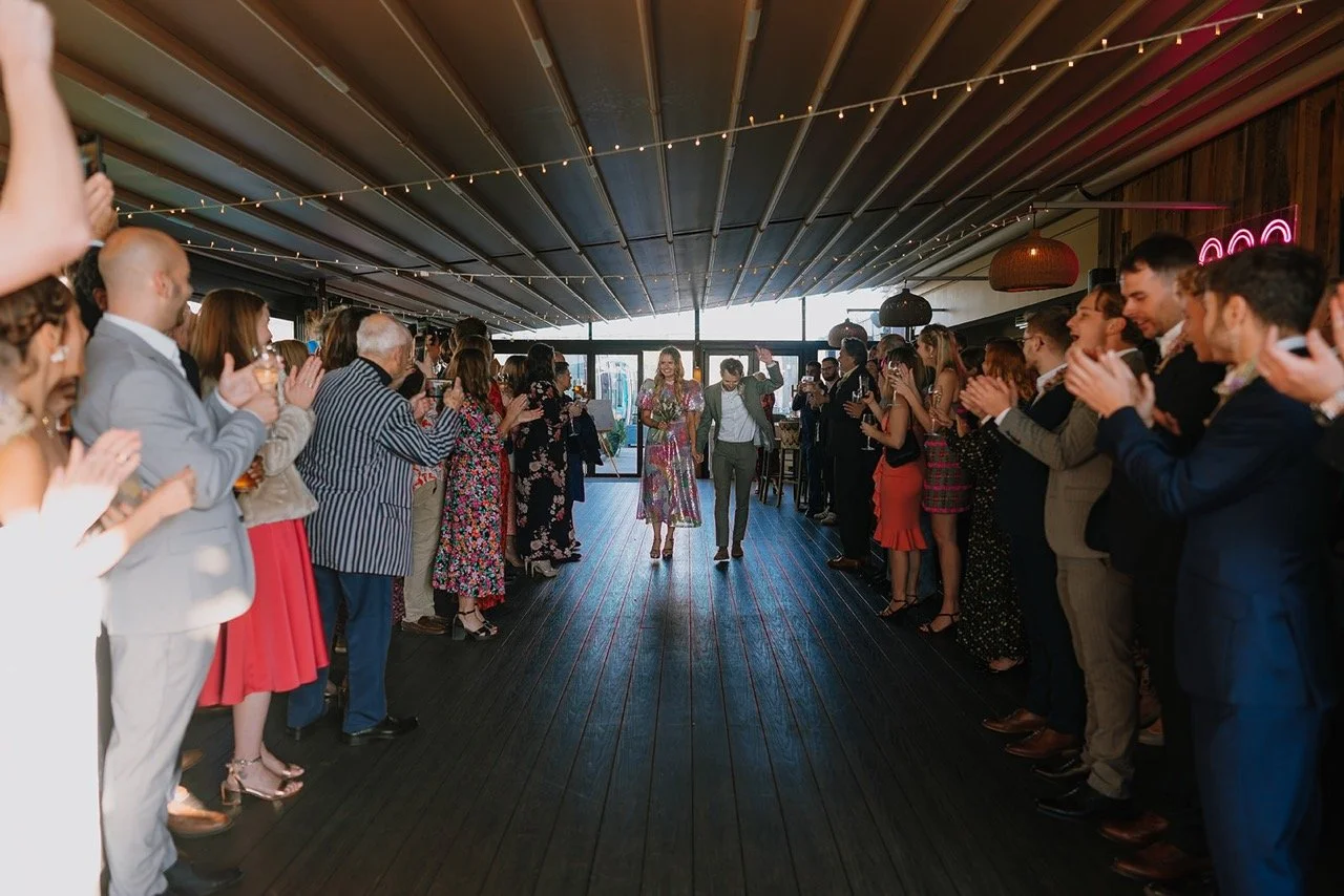 People clapping and cheering as a couple walks down the center of a decorated indoor venue during a celebration, possibly a wedding reception.