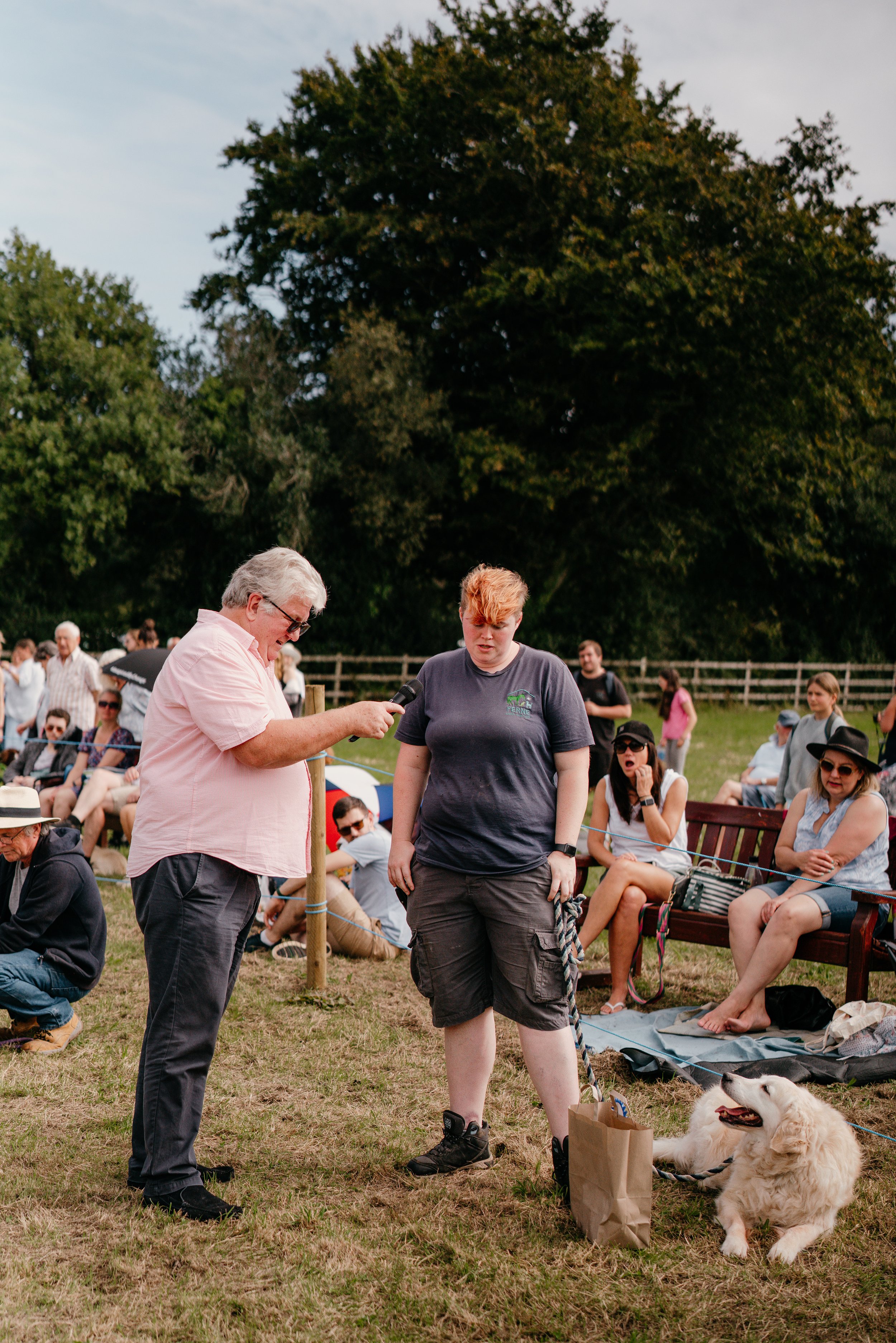 A man with gray hair and glasses is speaking into a microphone held by a young woman with short red hair, standing with a leash attached to her dog, a golden retriever, at an outdoor event with an audience seated on benches.