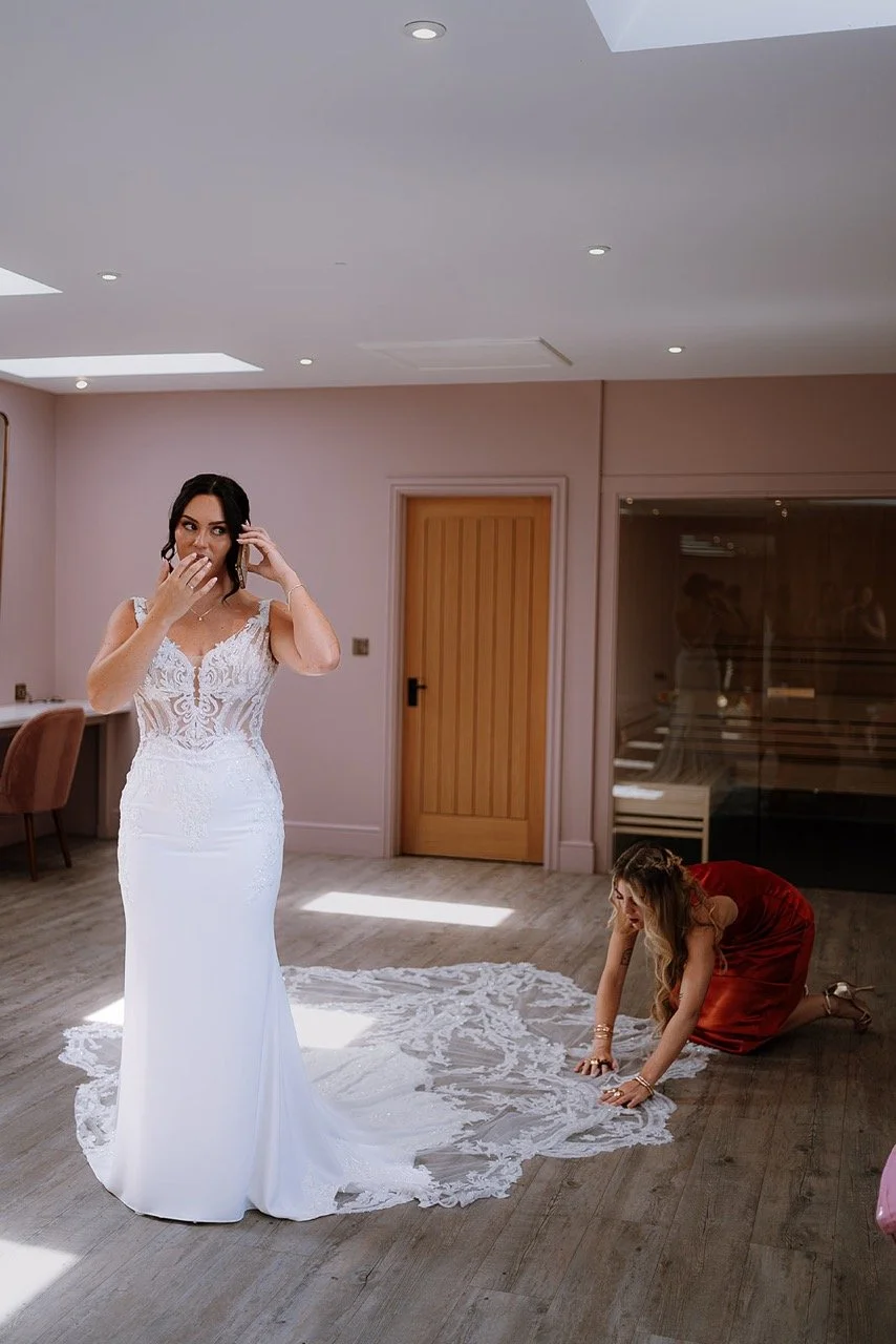 A woman in a white wedding dress standing in a room with two other women, one of whom is kneeling on the floor adjusting the train of the dress. The room has light pink walls, wooden doors, and a reflection of the women in glass.