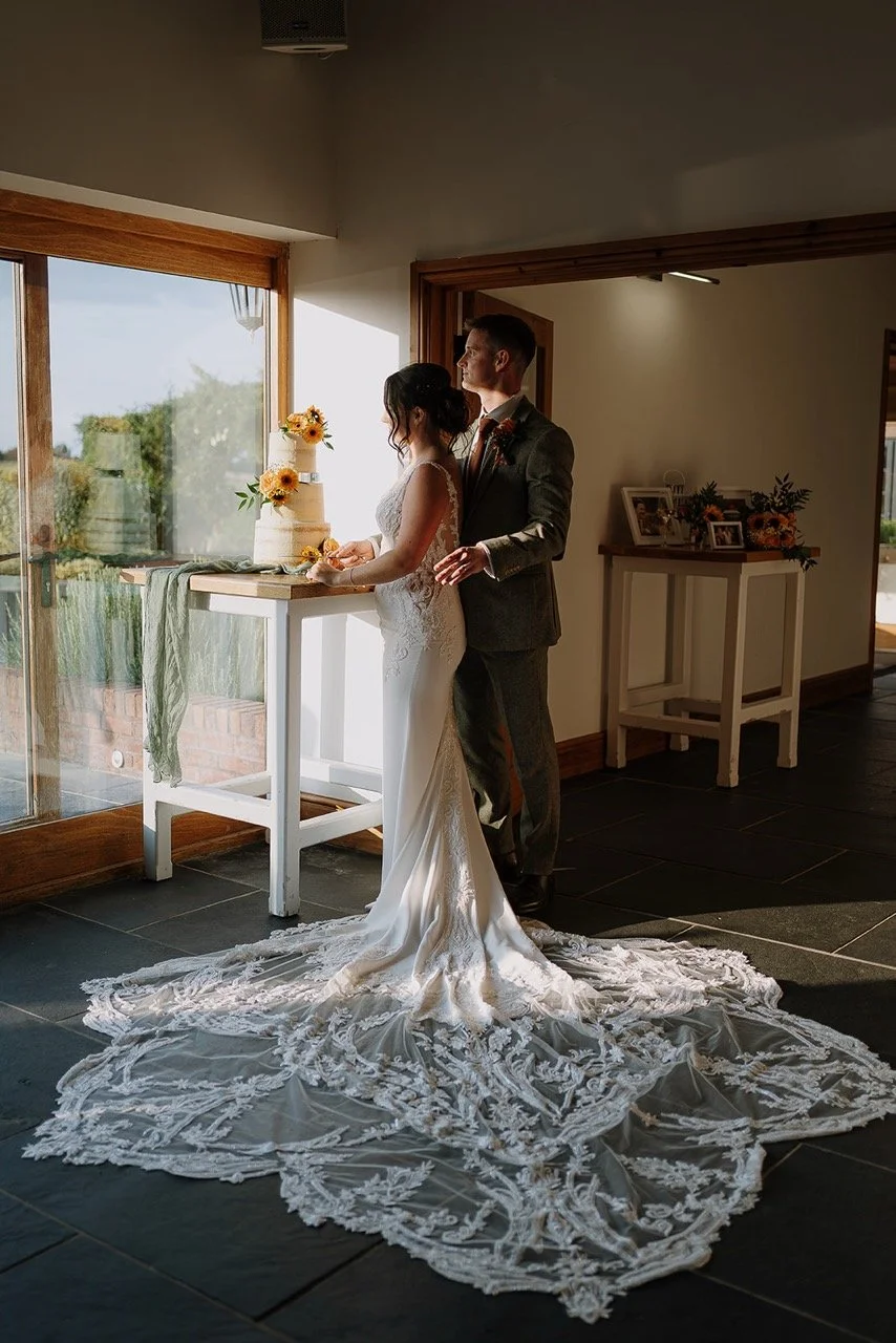 Bride and groom standing together in a room with sunlight coming through a window, near a wedding cake decorated with sunflowers.