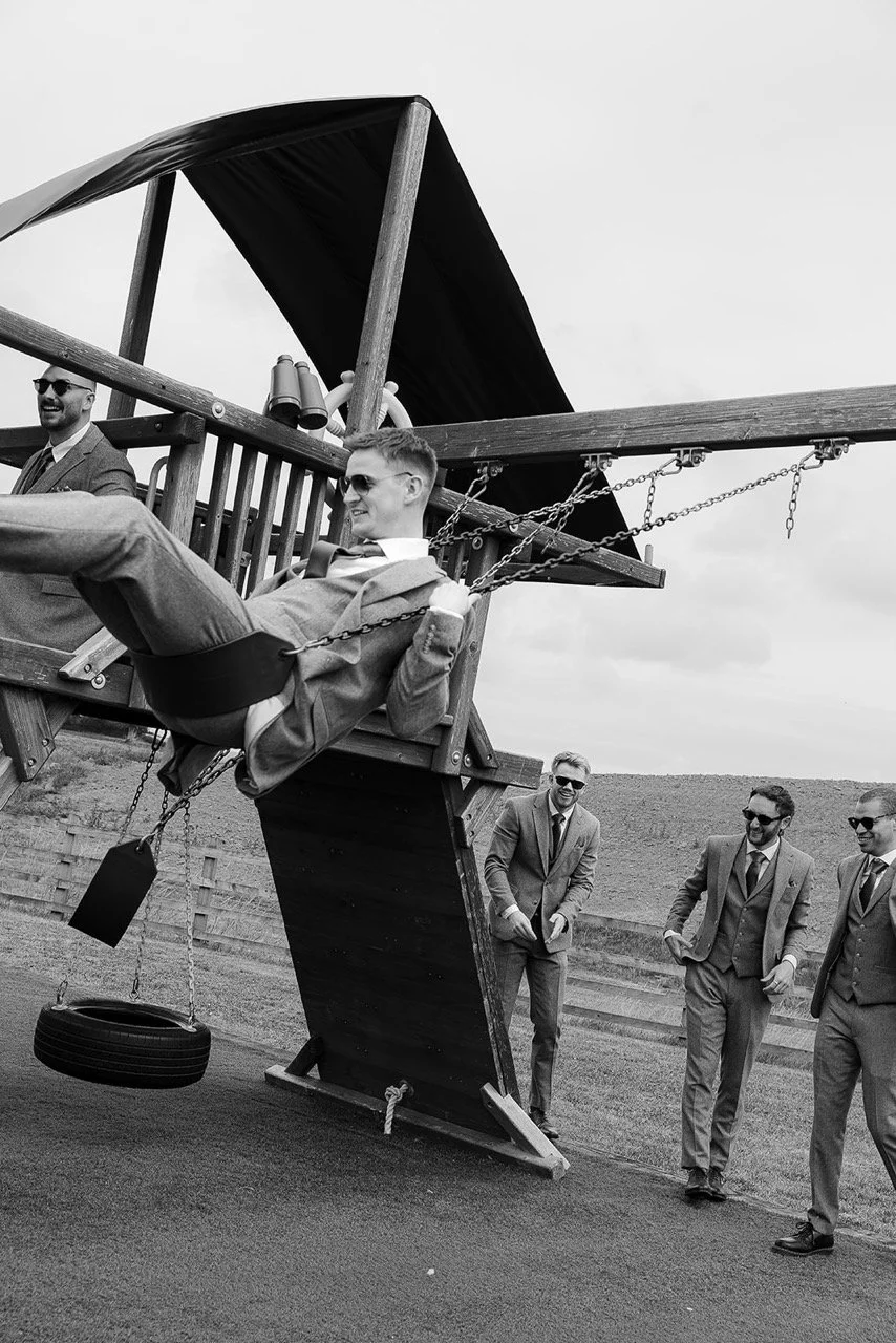 Black and white photo of four men in suits, three standing and one sitting on a swing attached to a wooden play structure resembling a ship, outdoors near a grassy field.