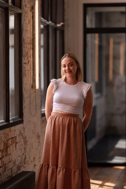 A woman standing indoors near a brick wall and window, smiling at the camera, wearing a white sleeveless top and a long brown patterned skirt.