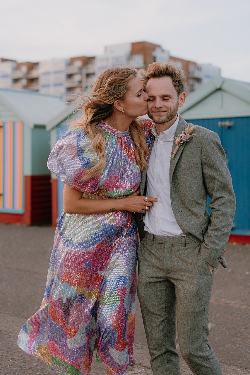 A woman kisses a man on the cheek at an outdoor location, with colorful beach huts and apartment buildings in the background. The woman is wearing a colorful, sequined dress and the man is dressed in a gray suit with a white shirt, smiling with close