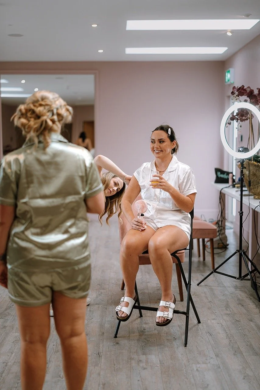 A woman sits on a chair, smiling and holding a drink while getting her hair styled by another woman in a salon or dressing room. The sitting woman is dressed in a white satin robe and sandals, with a hair clip and a small handheld fan. The stylist is