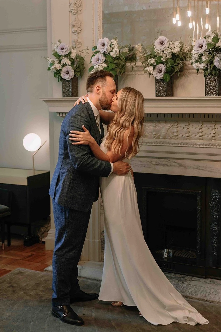 A couple in wedding attire sharing a kiss in a decorated indoor setting with flowers and a large mirror.