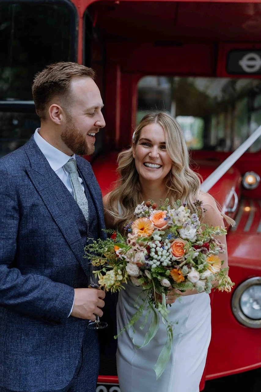 A smiling bride and groom stand together in front of a red vintage fire truck, with the bride holding a large bouquet of colorful flowers.
