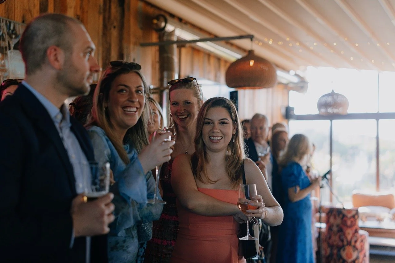 Group of people smiling and holding drinks at a social gathering inside a wooden venue with large windows and hanging light fixtures.