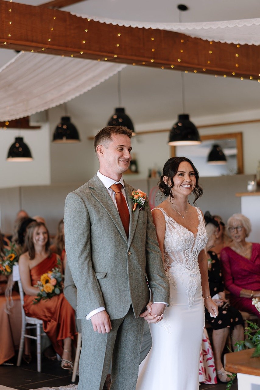Bride and groom holding hands during their wedding ceremony, smiling, with guests seated behind them in a decorated indoor venue.