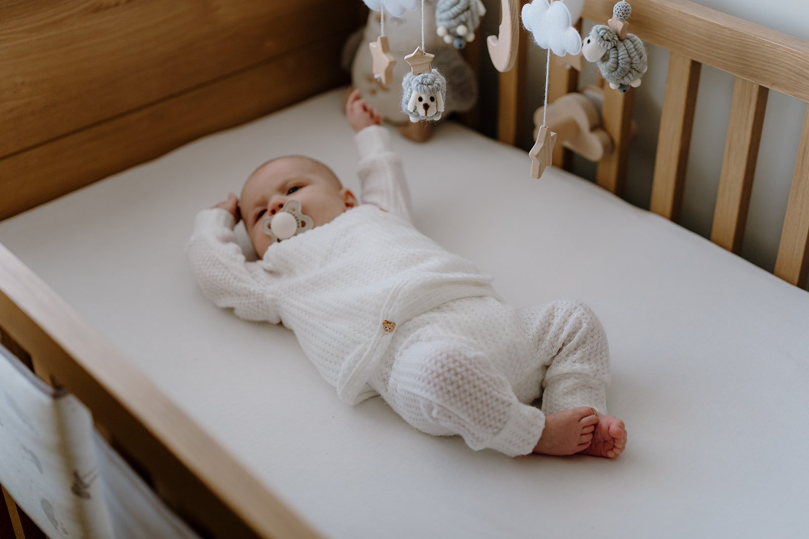 A baby lying in a crib with a mitten pacifier, wearing a white knitted outfit and surrounded by hanging plush toys and decorations in neutral tones.