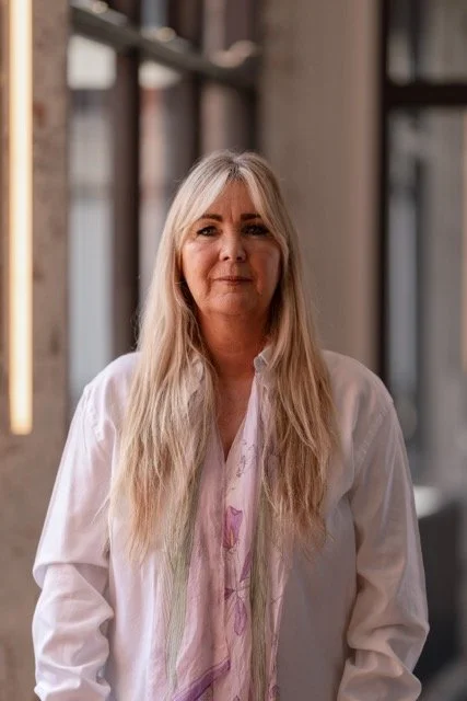 Woman with long blonde hair wearing a white blouse, standing indoors with blurred shelves and windows in the background.