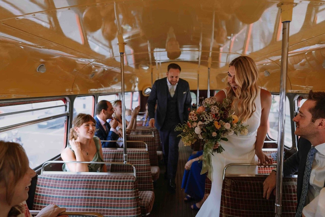 Bride in wedding dress holding a bouquet inside a vintage bus, smiling and surrounded by friends and family.
