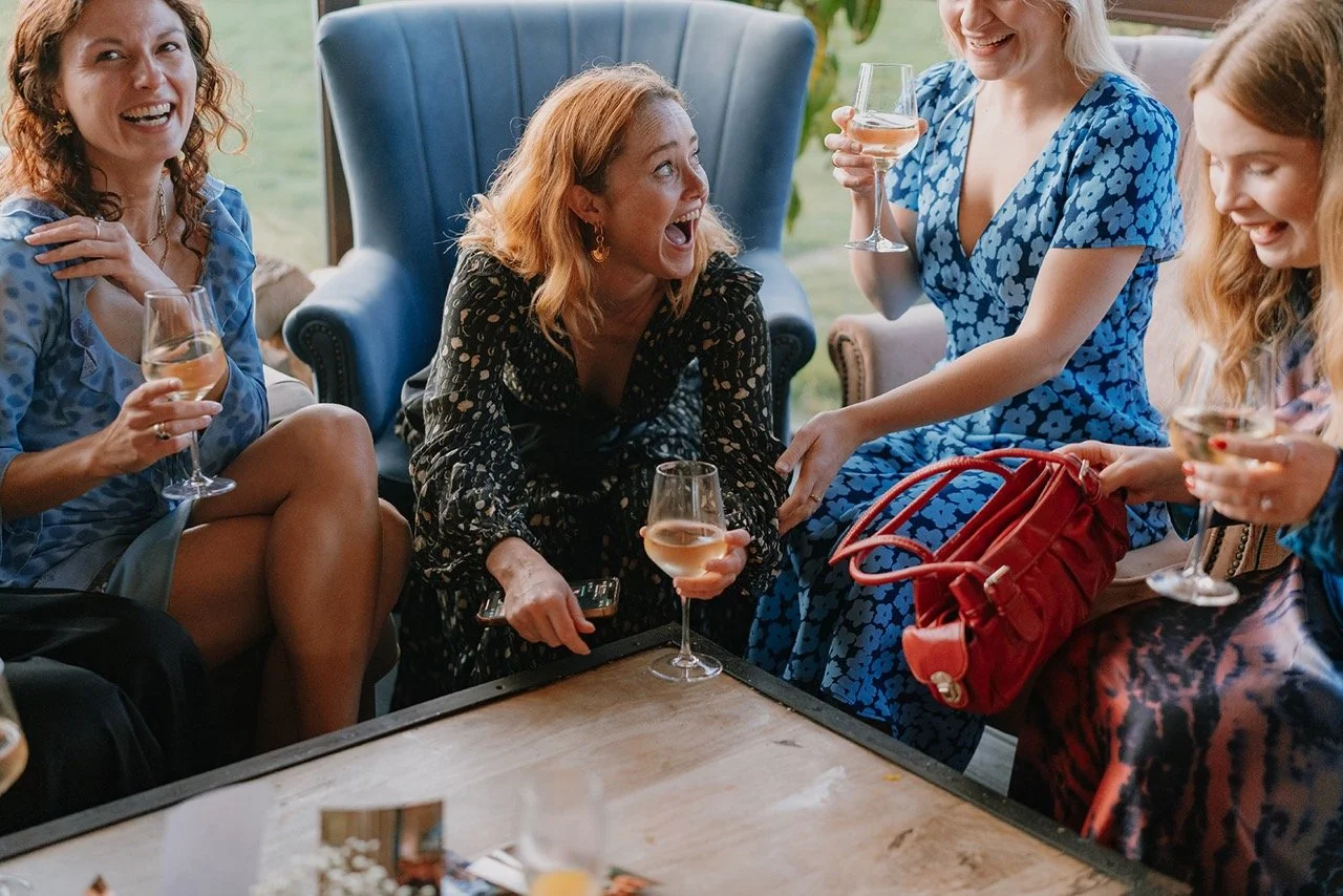 A group of women laughing and socializing while holding wine glasses in a cozy indoor setting.