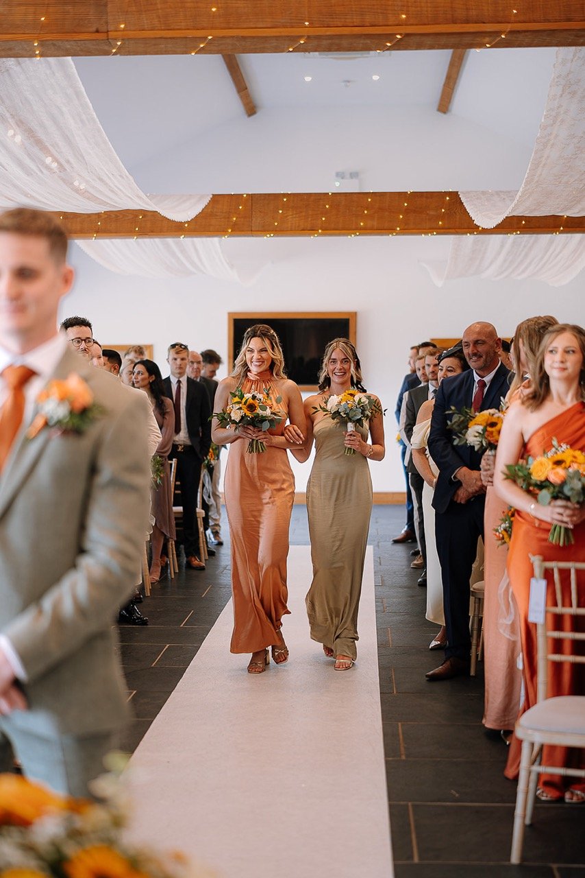 Two women walking down an aisle at a wedding, holding bouquets, smiling, with guests seated on either side.