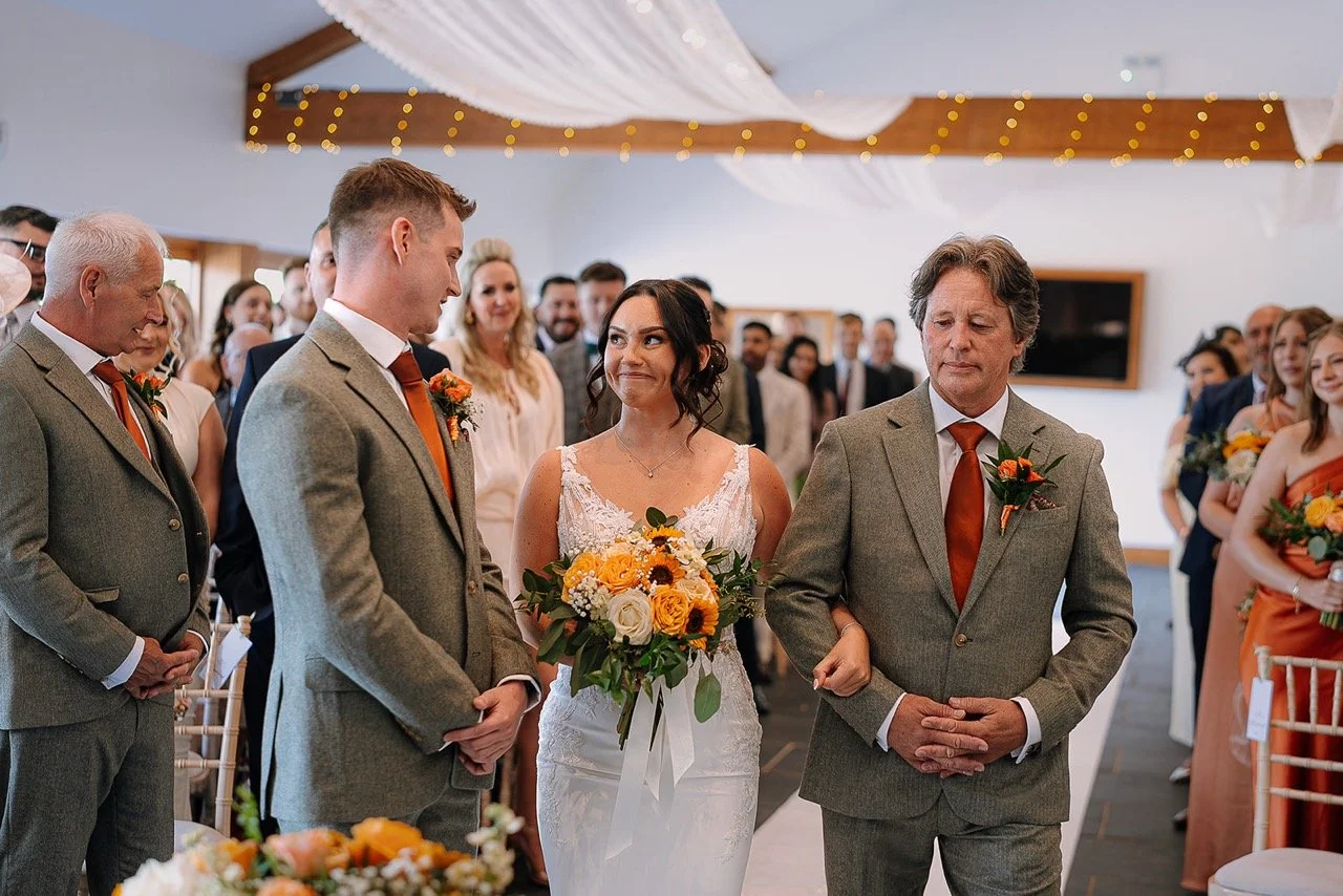 Bride walking down aisle with her father, surrounded by wedding guests, at a wedding ceremony indoors