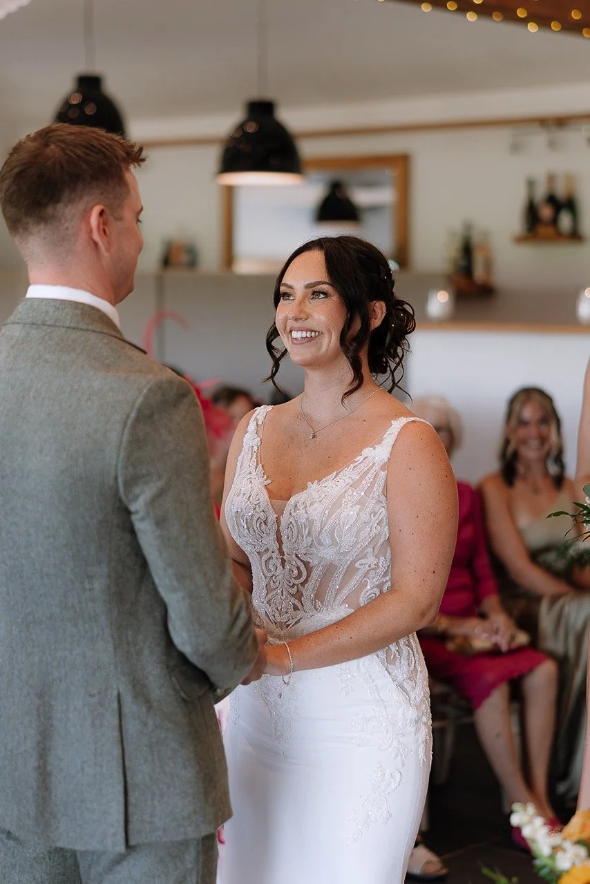 A bride and groom exchanging vows at their wedding indoors, with the bride wearing a white lace wedding gown and the groom in a grey suit, surrounded by seated guests.