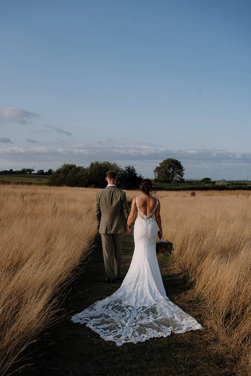 A bride and groom walking hand-in-hand along a narrow path through tall, golden grass in a rural landscape under a blue sky with some clouds.
