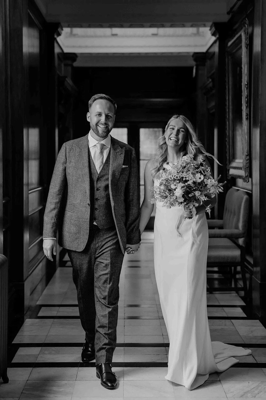 A black and white photo of a bride and groom walking hand in hand inside a historic building, smiling, with the bride holding a large bouquet of flowers.