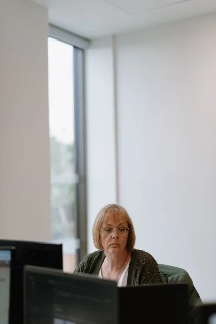 An elderly woman with glasses and short hair sitting at a desk in an office, looking at a computer screen, with a large window in the background.