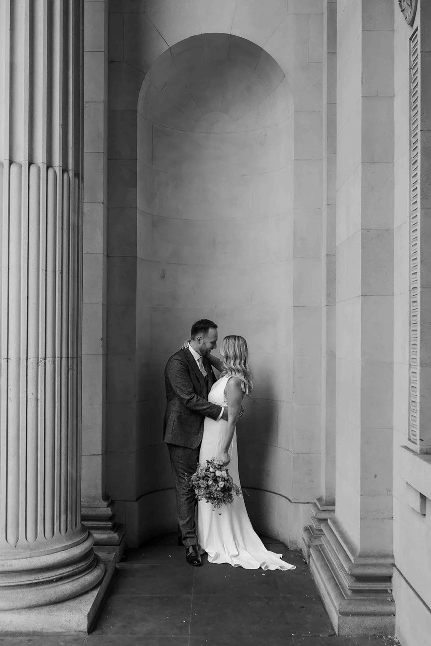 A black and white photo of a couple on their wedding day standing close together, with the man touching the woman's shoulder, in front of a large stone wall with columns.