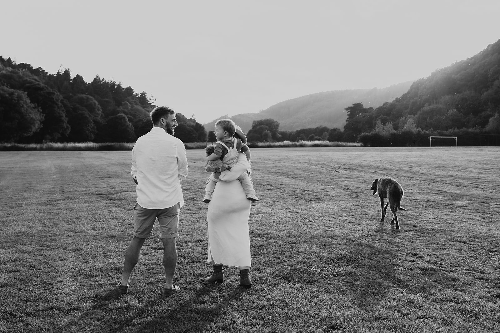 A black-and-white photo of a family on a grassy field, with a man, a woman holding a child, and a dog walking in the background, with hills and trees in the distance.