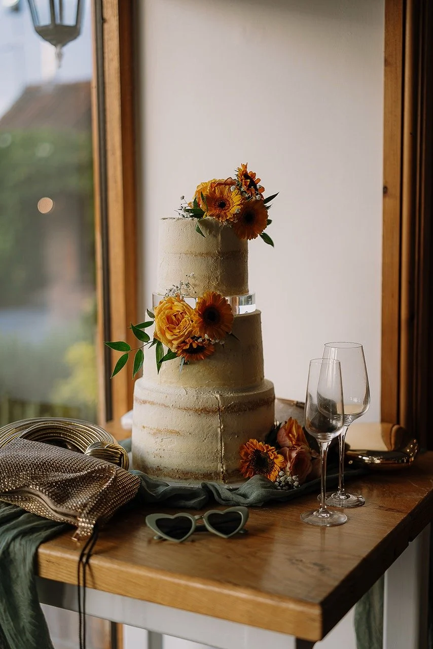 A three-tiered wedding cake decorated with orange and yellow flowers, placed on a wooden table with glasses and accessories nearby, near a window with a view outside.