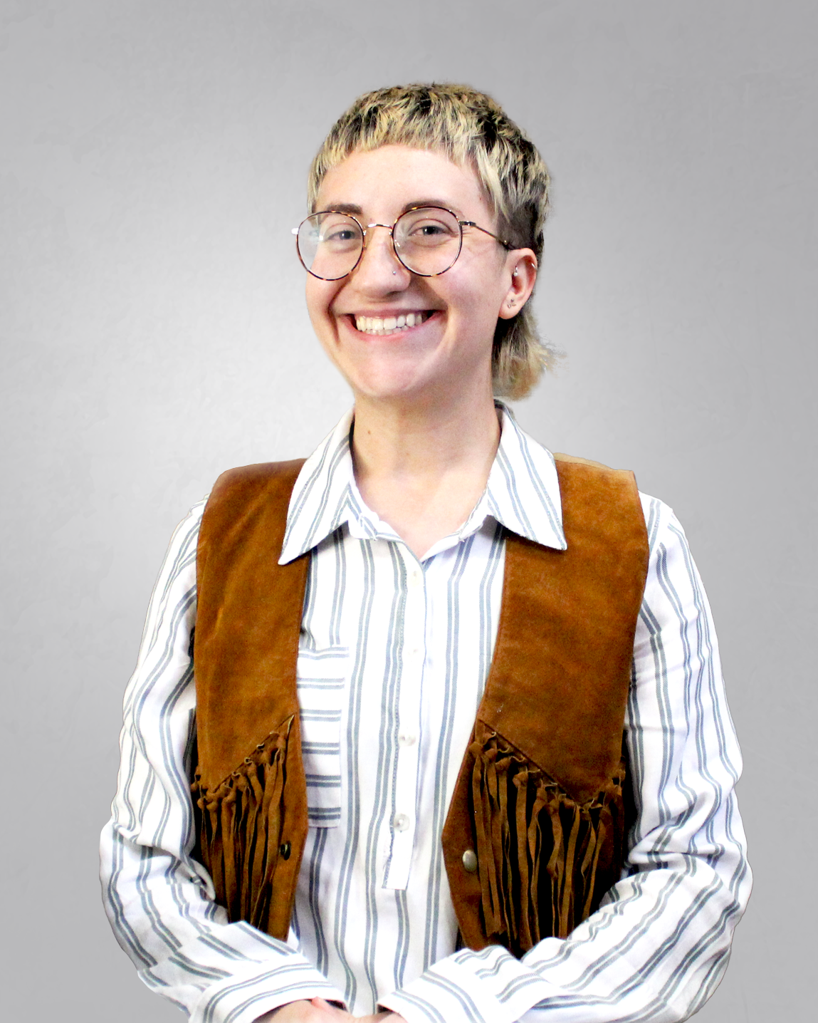 Smiling woman with short blond hair, glasses, wearing a striped shirt and fringe leather vest, sitting in front of a multicolored brick wall.