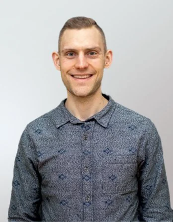 Portrait of a young man with short hair, smiling, wearing a patterned button-up shirt against a dark background.