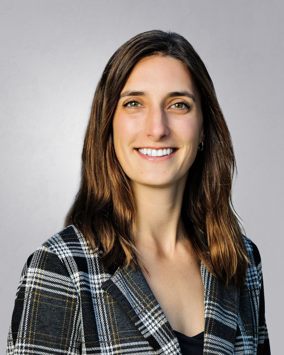 Portrait of a smiling woman with shoulder-length brown hair, wearing a black and white plaid blazer, standing outdoors in front of a rocky background.
