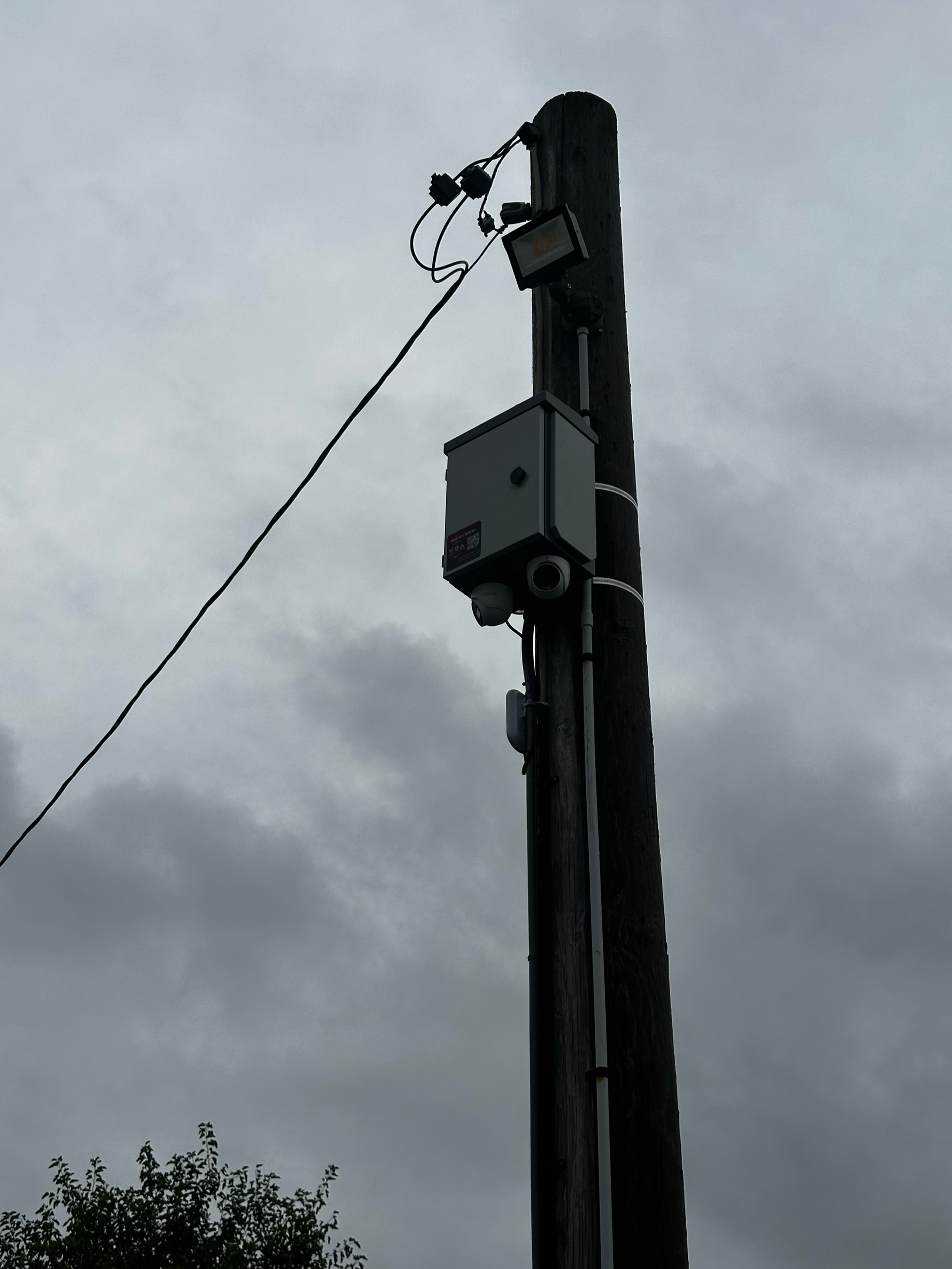 Close-up of a utility pole with electrical equipment, a security camera, and a floodlight, against a cloudy sky.