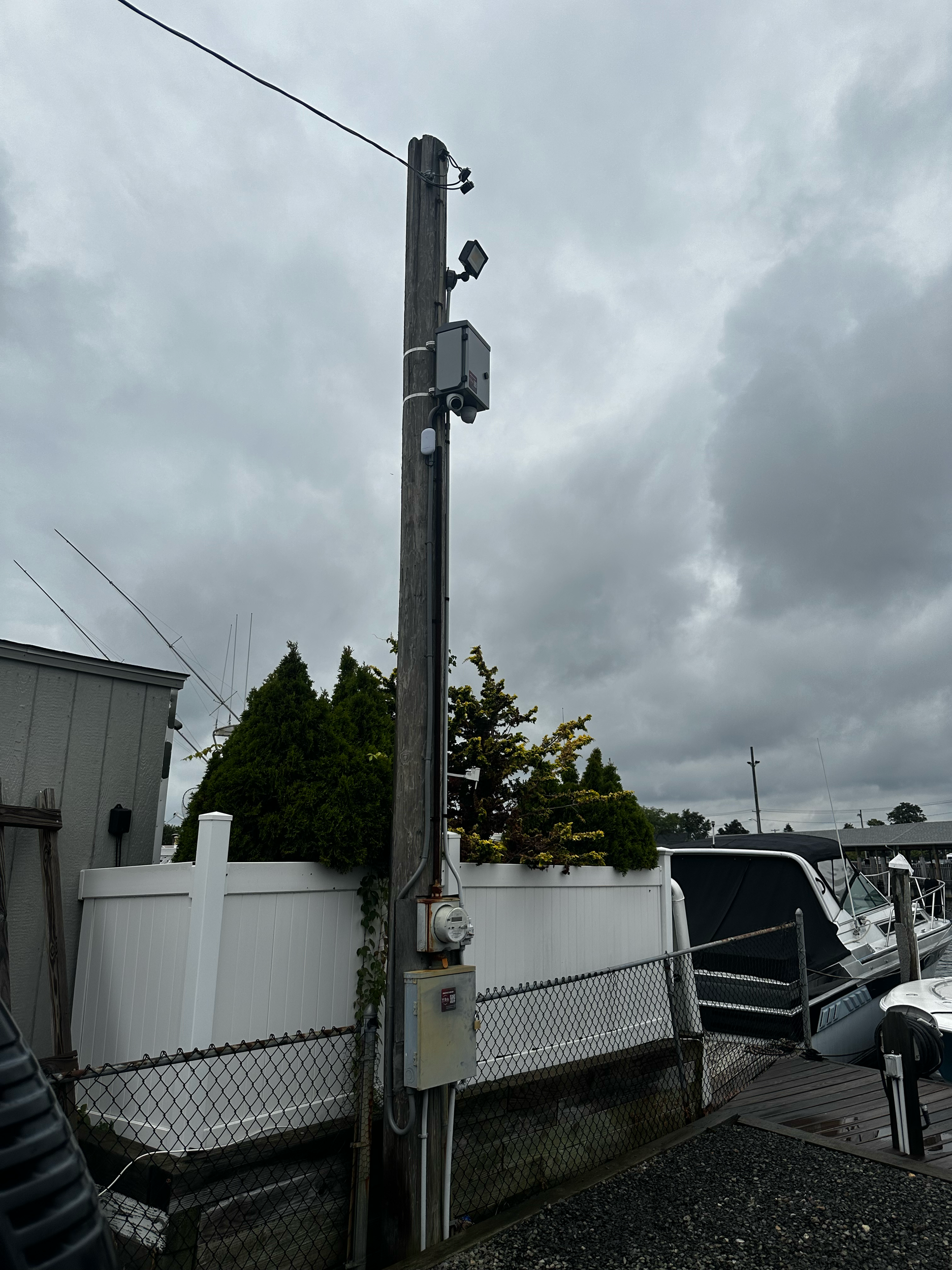 A utility pole with surveillance cameras and electrical equipment, situated next to a boat docked in a marina with a cloudy sky overhead.