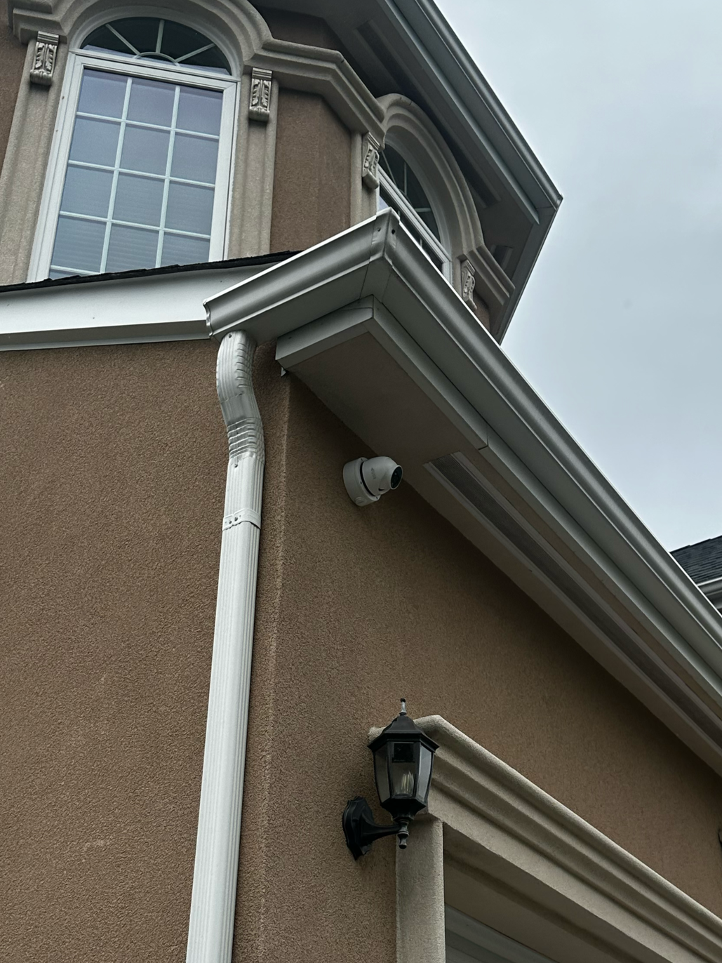 Close-up of the corner of a house showing a beige textured exterior wall, a white gutter system, a surveillance camera, and a decorative outdoor wall lantern.