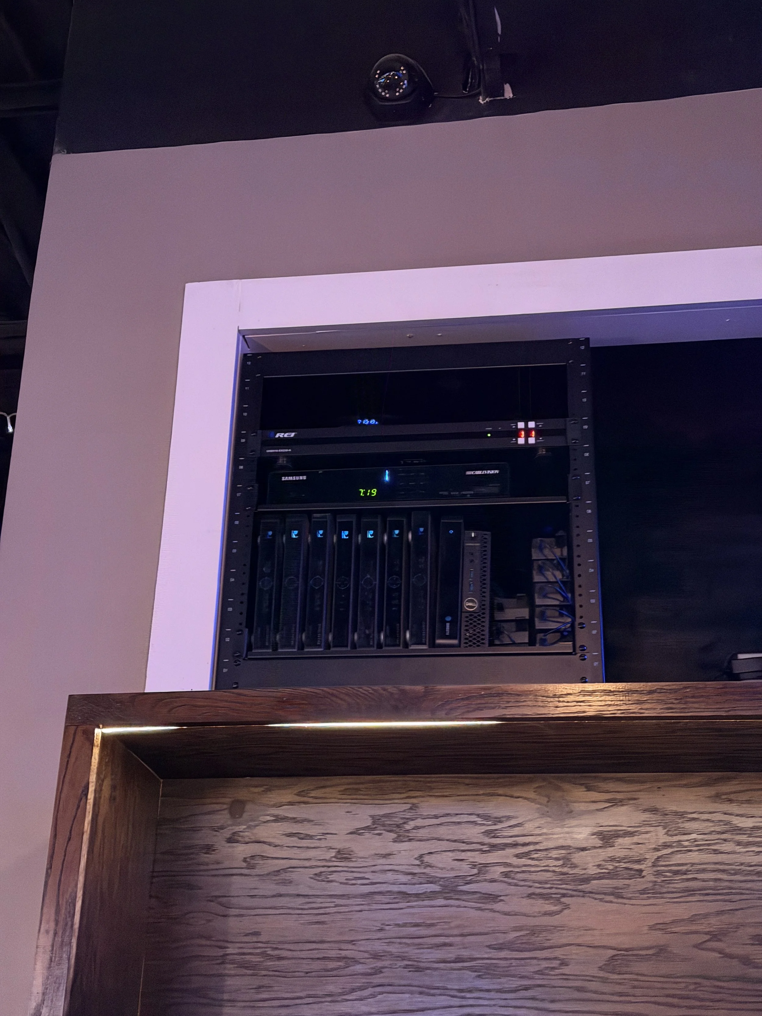 A server rack with multiple servers and networking equipment, visible on a dark wall, with a wooden surface in the foreground.
