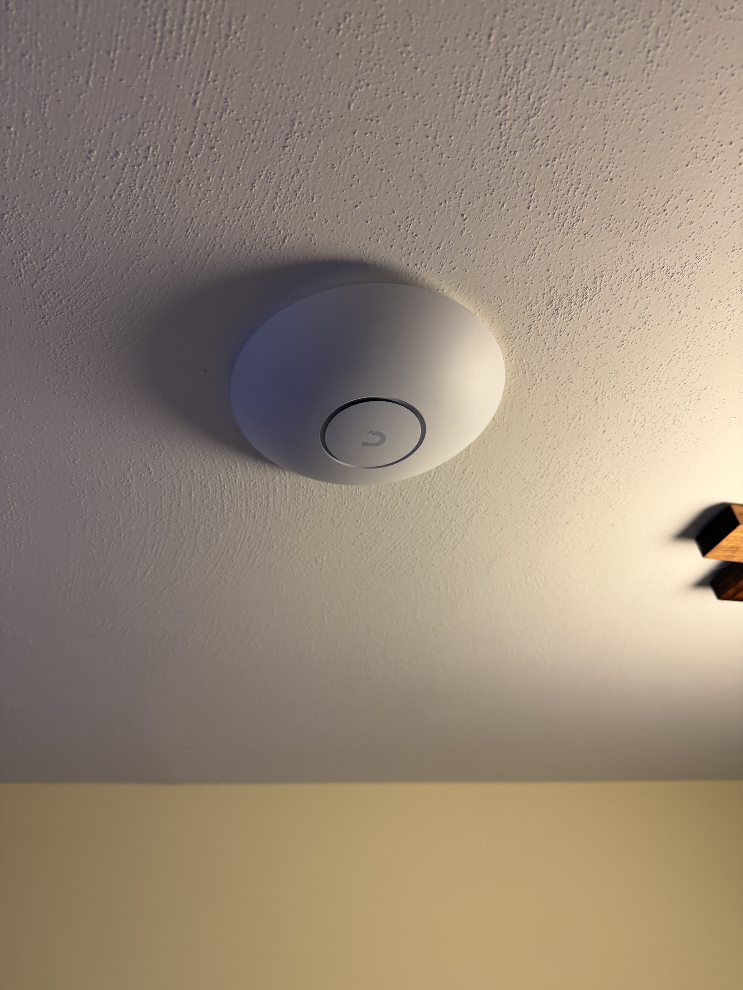 Ceiling-mounted smoke detector with a circular design and a power button in the center, attached to a textured ceiling. Part of a wooden ceiling beam is visible on the right side.