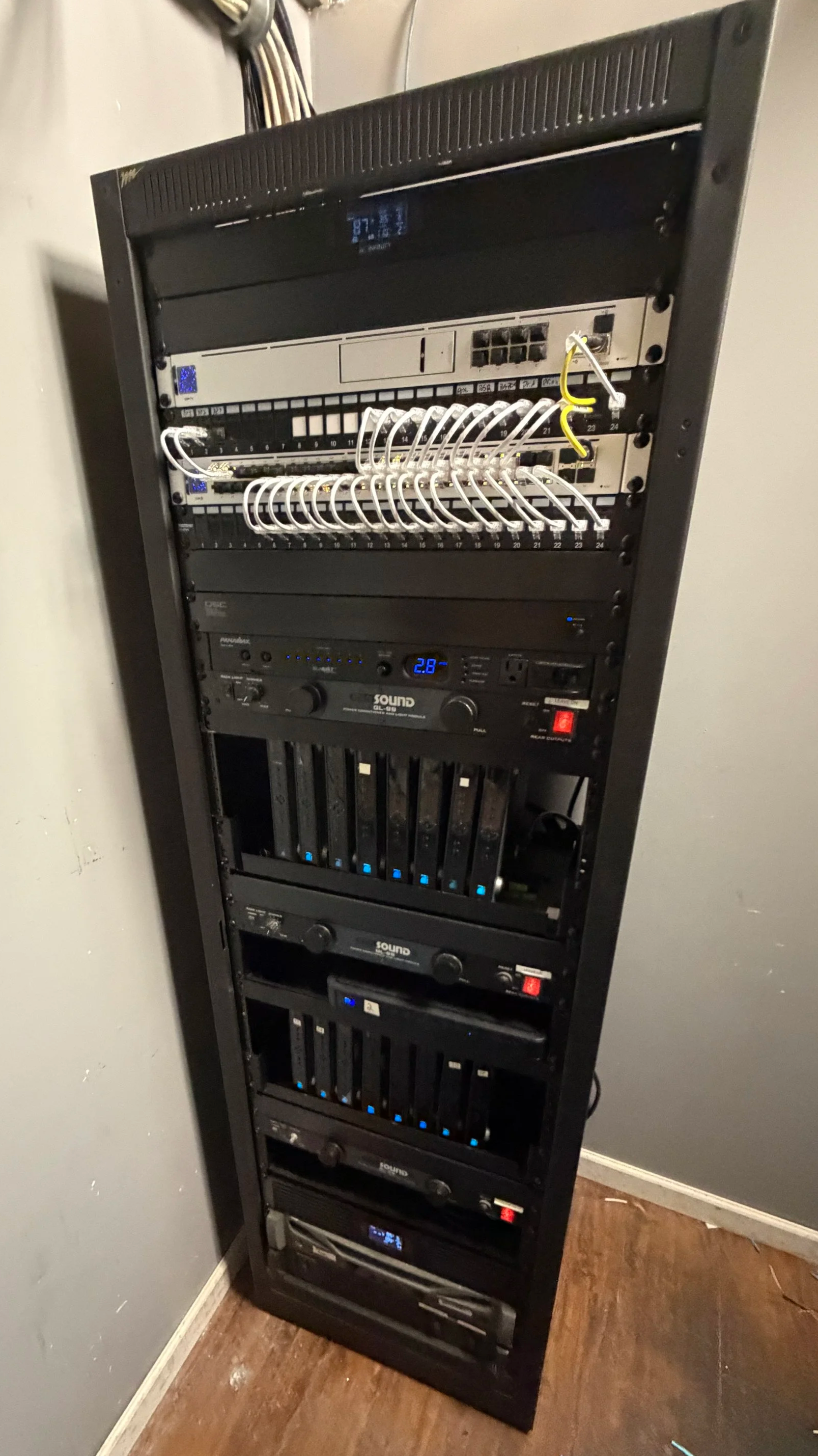 A tall black server rack containing networking and audio equipment, with various cables connected at the top, and blue indicator lights on some units.