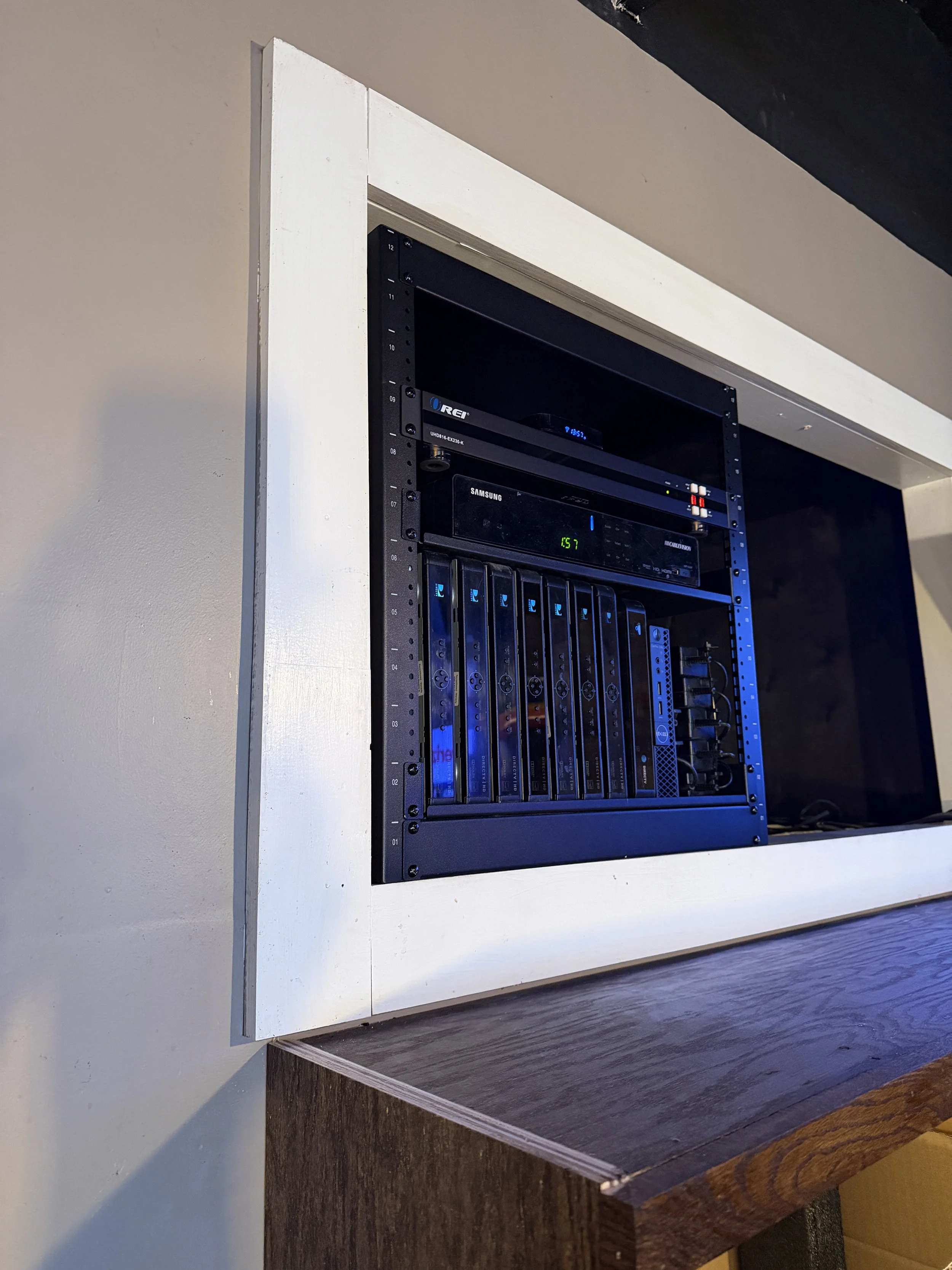 Open wall-mounted cabinet containing computer networking equipment including a router, switches, and a disk drive, set in a white frame with a dark wall behind.