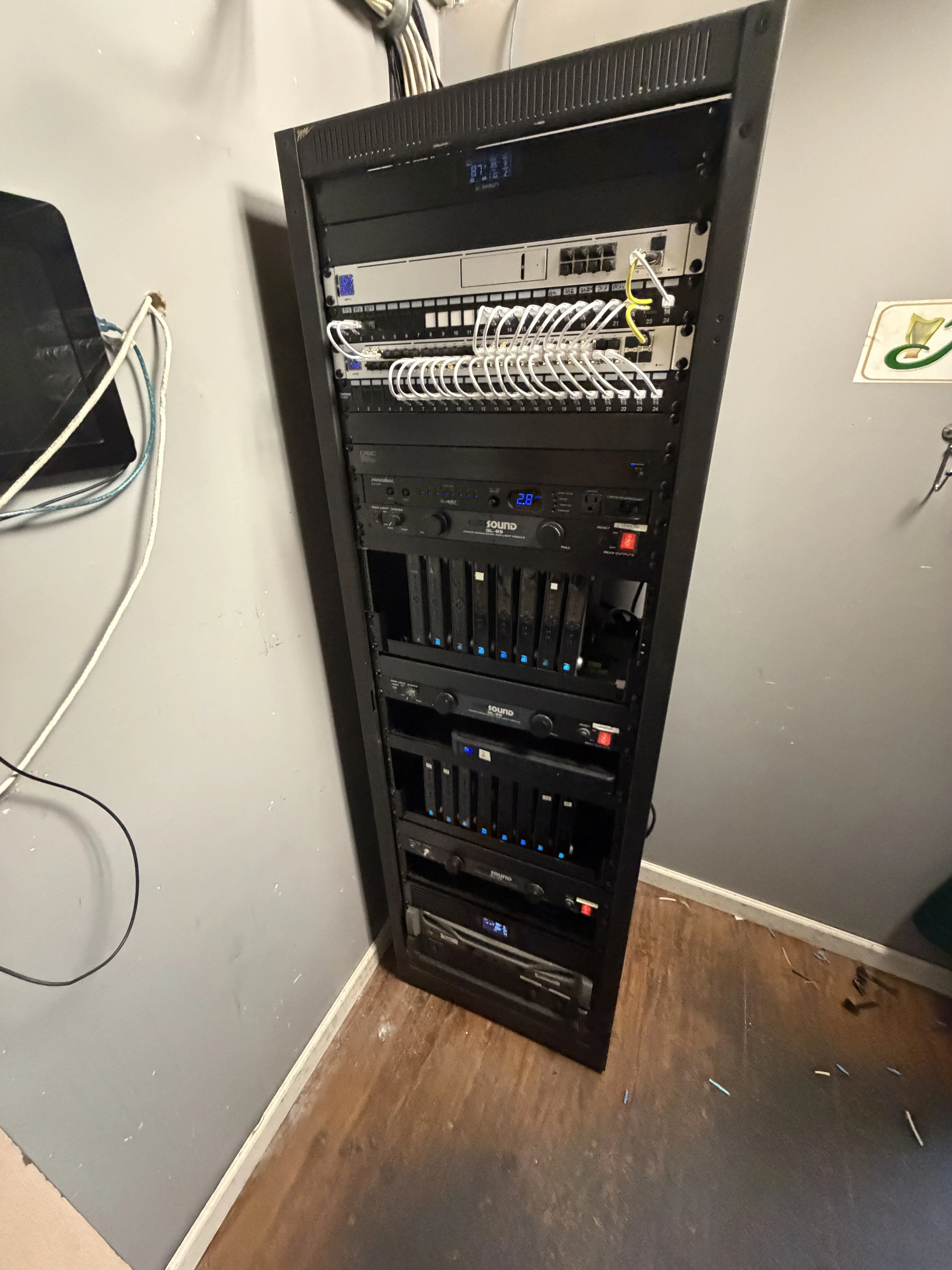 A tall black audio equipment rack with various electronic devices, including a sound system, audio amplifiers, and networking equipment, placed against a light gray wall with some wires and small objects on the floor nearby.