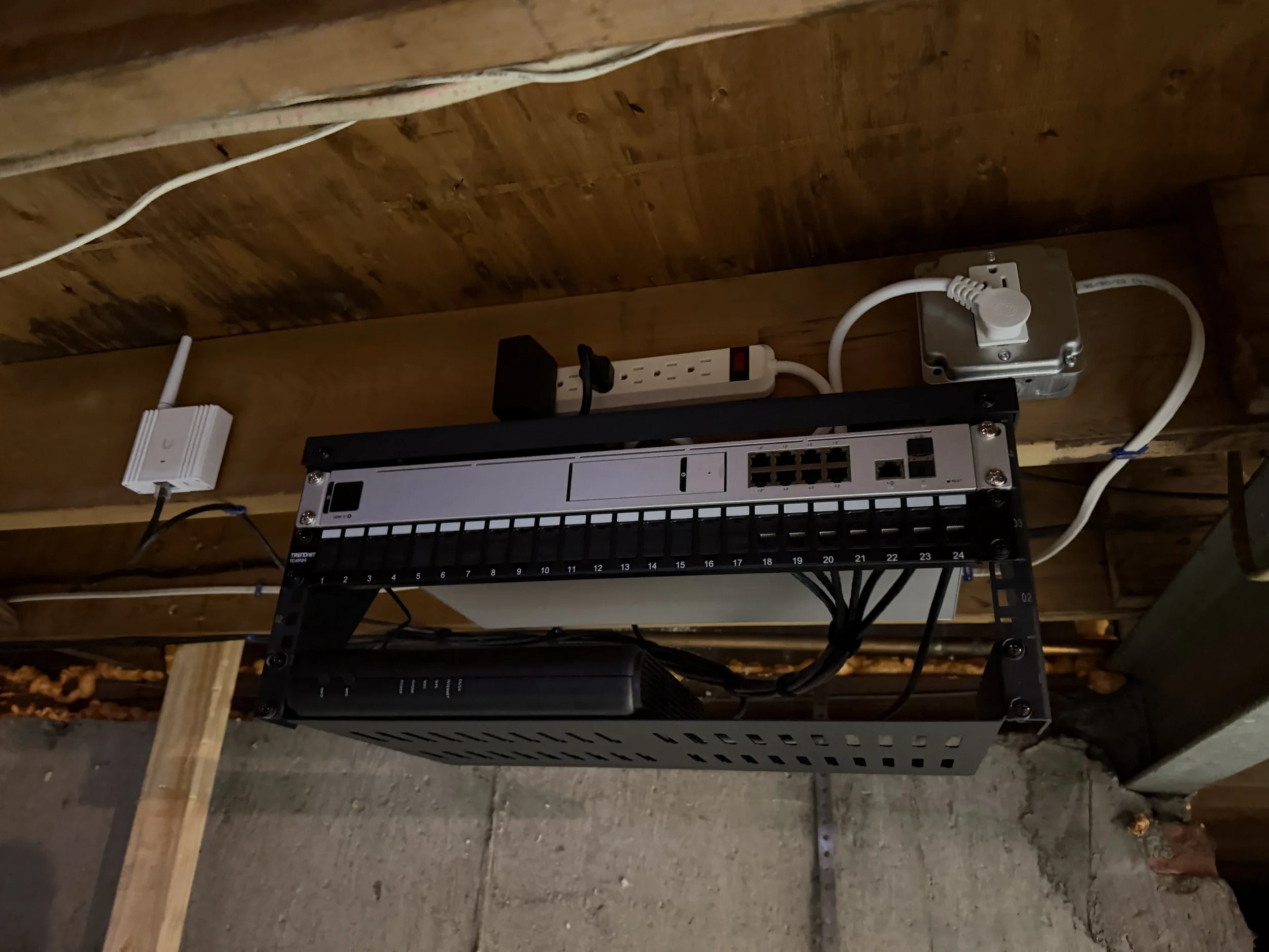 Electrical network equipment mounted under a wooden ceiling, including a power strip, a network switch, a device enclosure, and various cords and cables.
