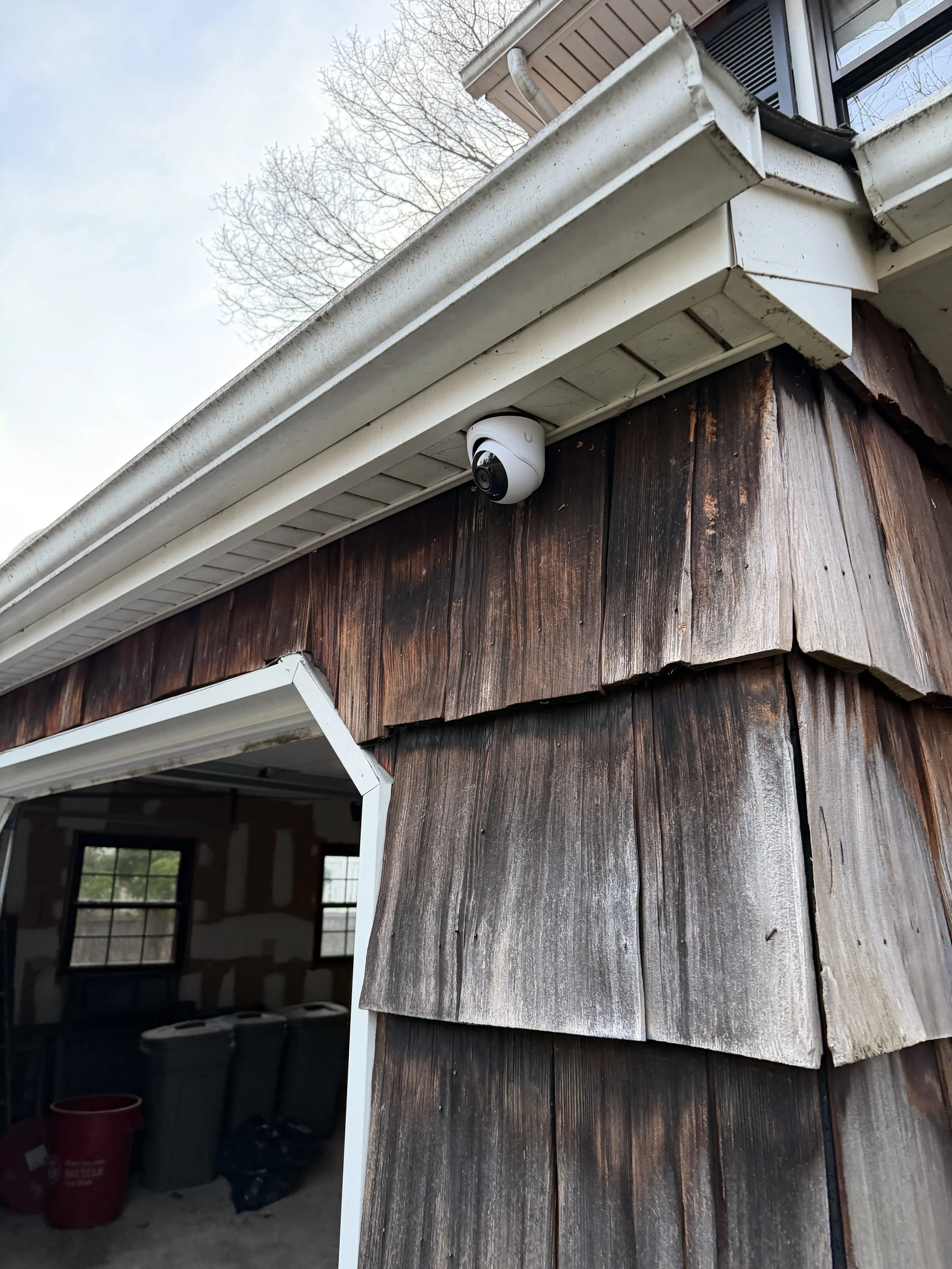 Close-up of a corner of a building with weathered wooden siding, a white security camera mounted under the eaves, and a partly open door revealing a cluttered interior with trash bins.