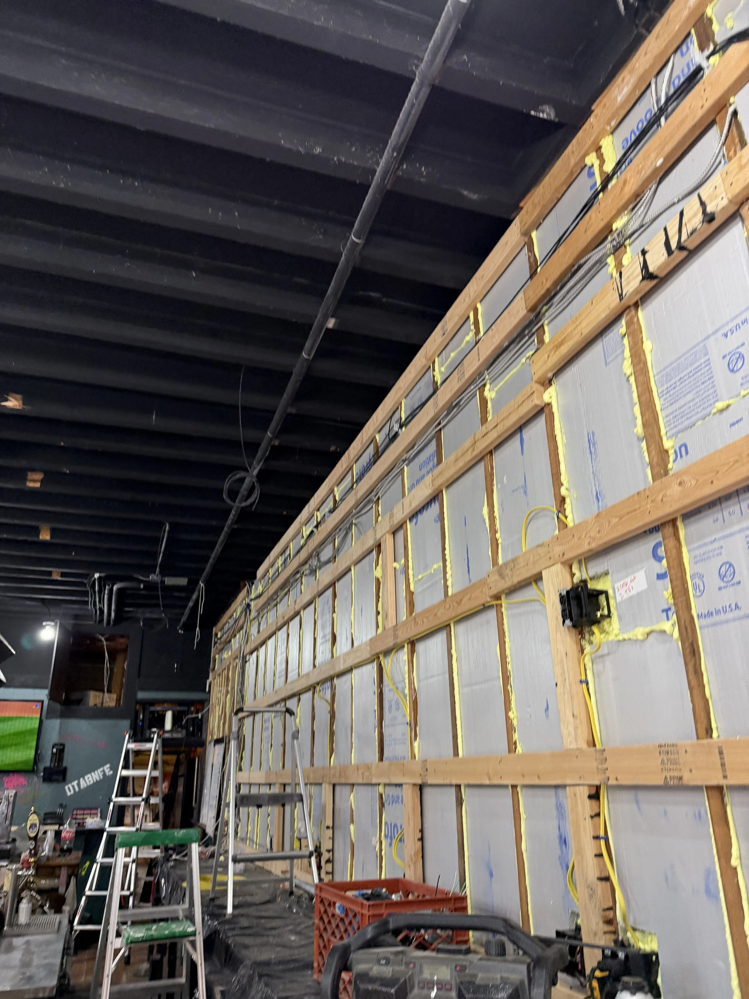 Interior of a building under construction with wooden framing, insulation, electrical wires, ladders, and construction tools.