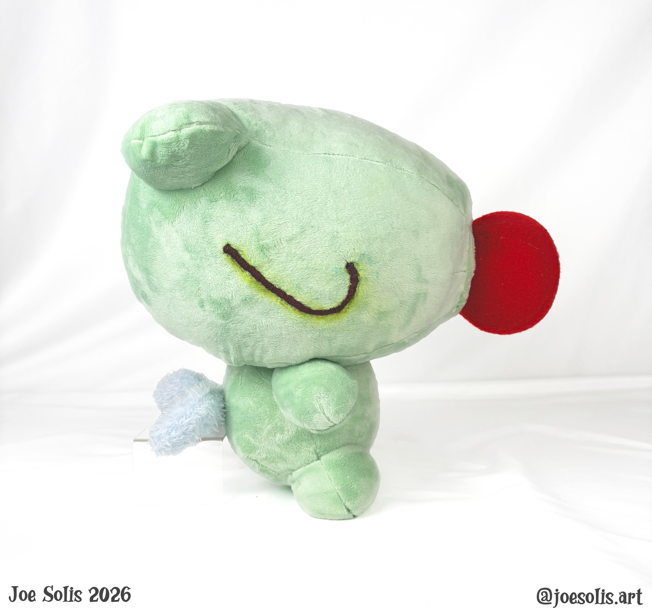 A plush toy of a green dinosaur with a red crest on its head, facing downward, with a white background and black text at the bottom.