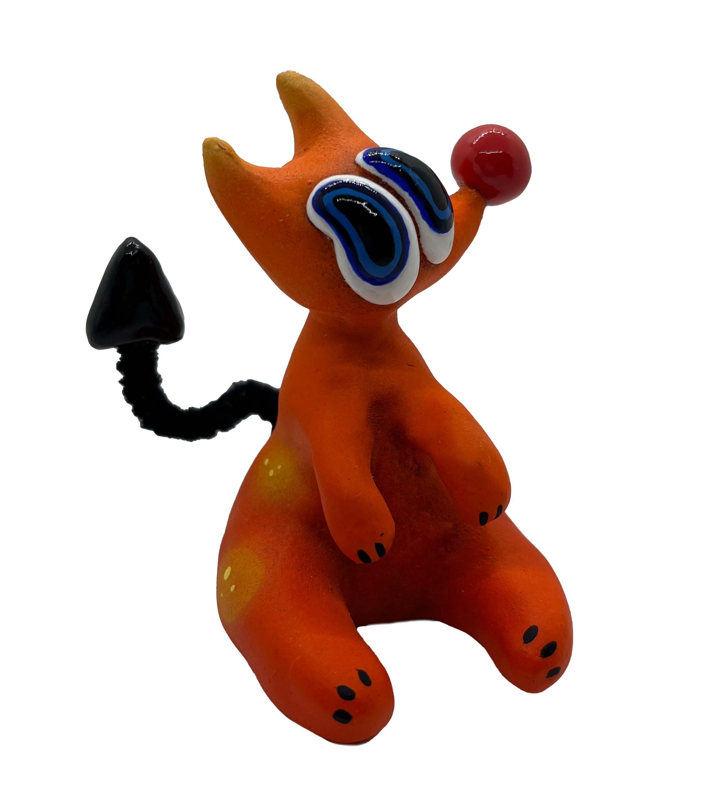 Orange figurine resembling a cat with large blue and black eyes, black dots on paws, and a red ball on its tail, with a black object partially visible at left.