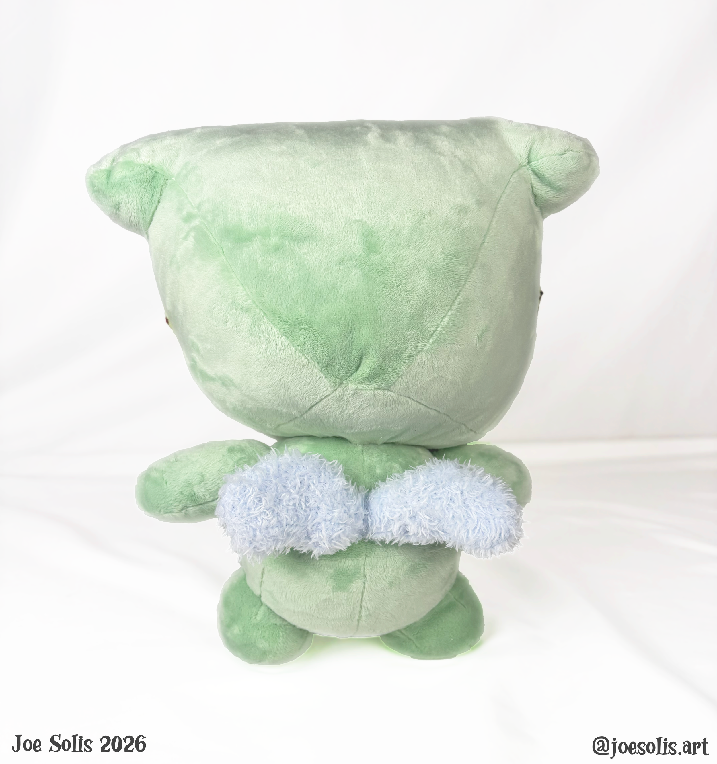 A pastel green stuffed animal, possibly a dinosaur, with a white fluffy patch on its belly, upside down against a white background.
