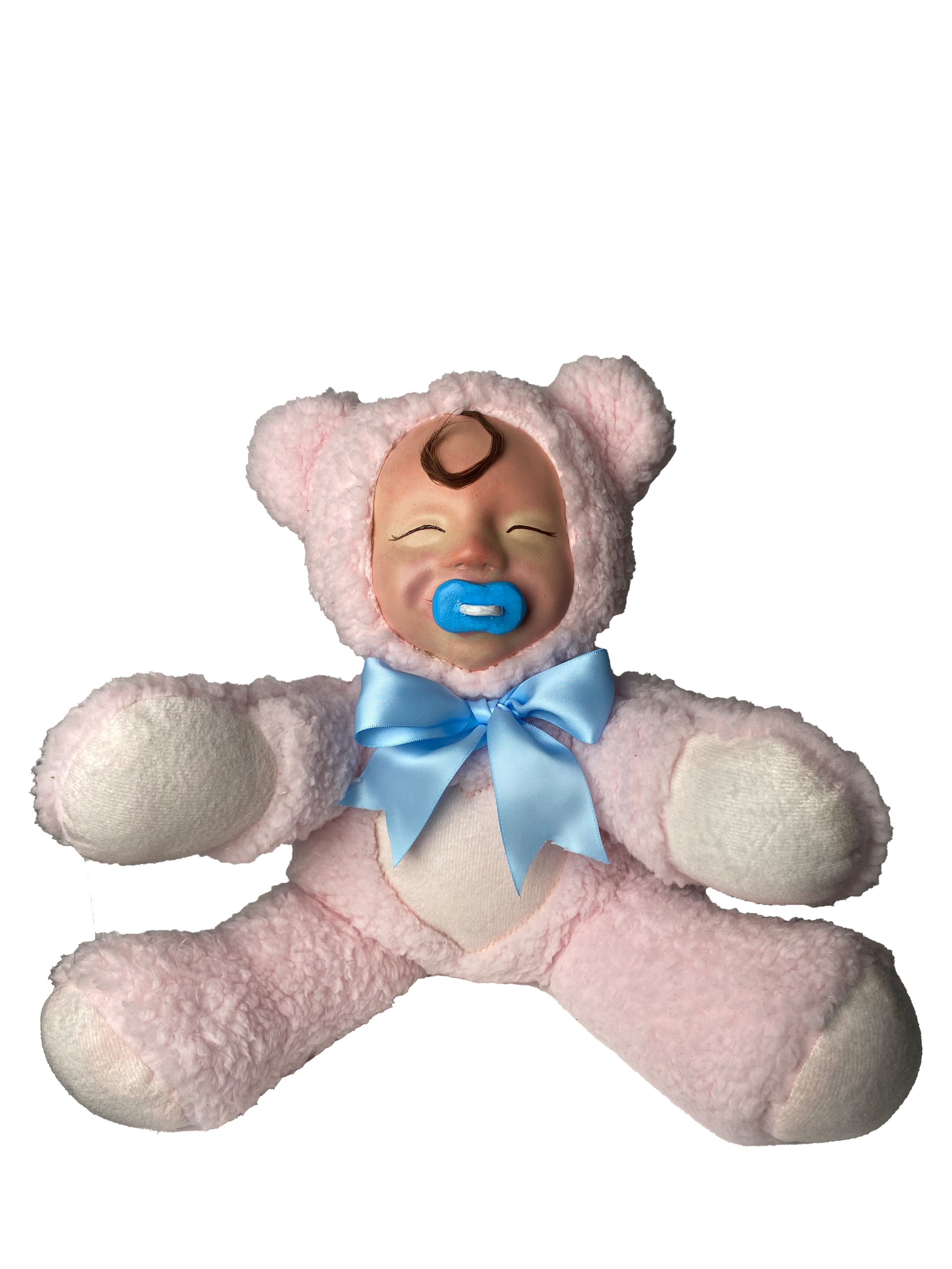 A plush teddy bear toy with a human baby doll face inside a pink teddy bear costume. The doll has closed eyes, a curl of hair on its forehead, and a blue pacifier in its mouth, with a blue ribbon tied around its neck.
