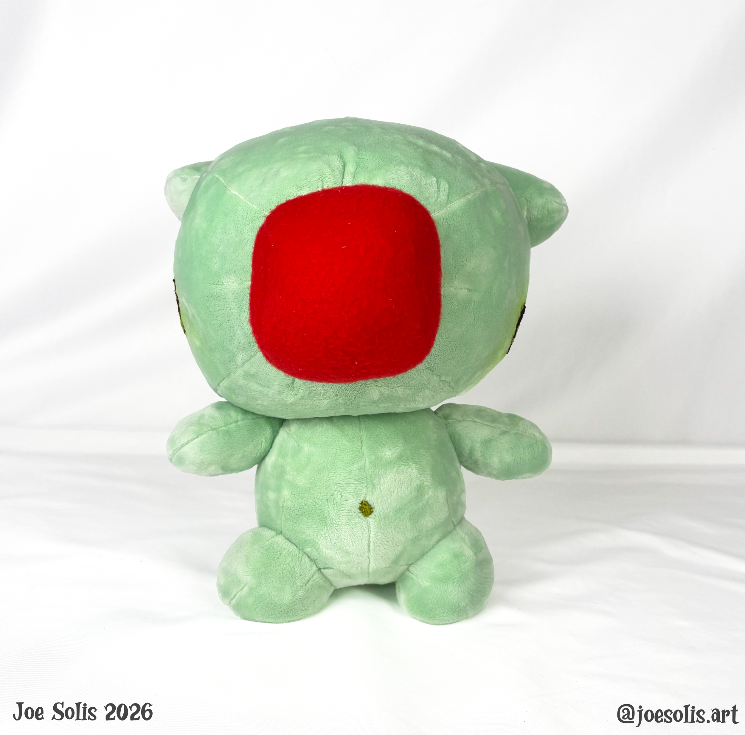 A plush toy of a green dinosaur with a large red nose, facing away from the camera, standing on a white surface.