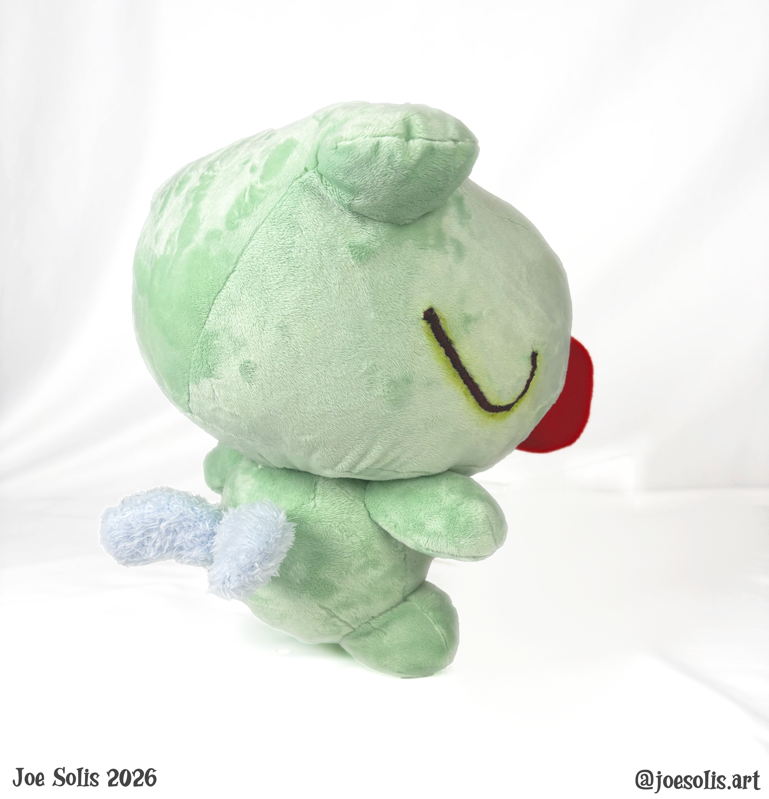 A close-up photo of a green plush stuffed animal with white paws, a rounded head with a folded ear, black embroidered eyes, and a black embroidered smiling mouth, possibly a frog or turtle, against a plain white background. The bottom left contains t