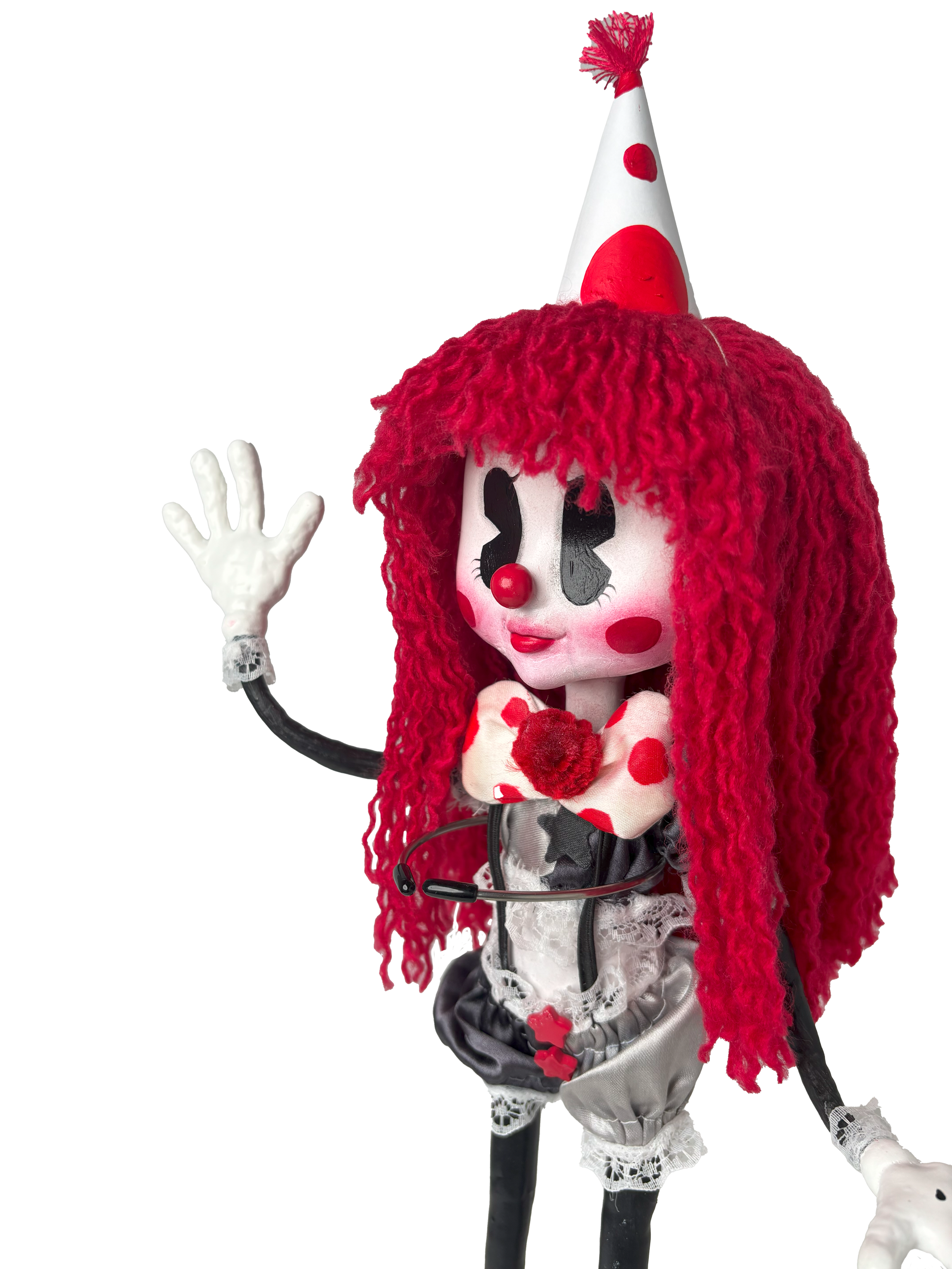 Close-up of a clown doll with exaggerated facial features, curly red hair, a white face with black eye makeup, a red nose, and red cheeks, wearing a cone-shaped party hat with red polka dots and a red pom-pom on top, a red and white polka-dotted bow 
