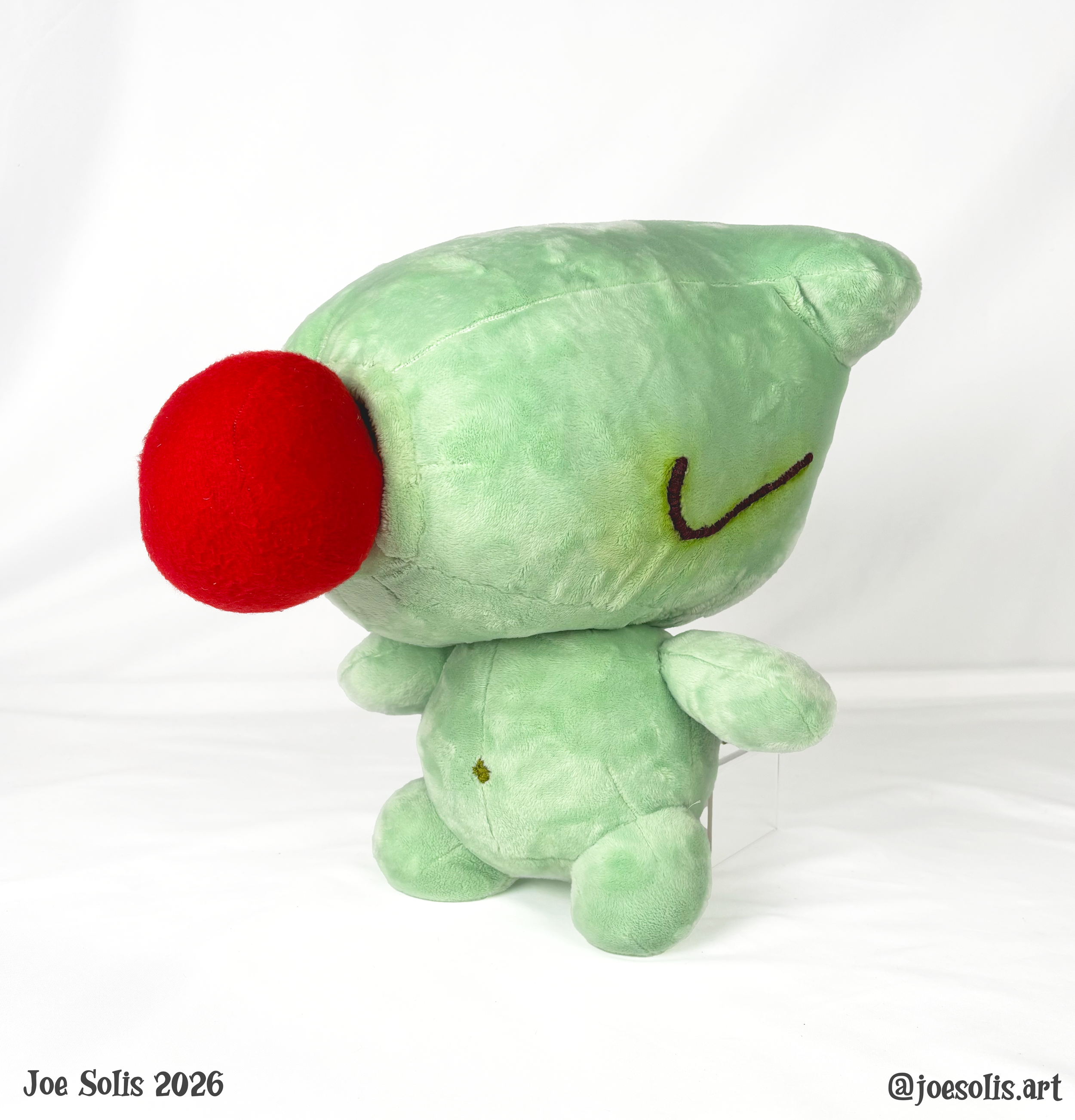 A plush toy frog with a red nose, closed eyes, and a smiling mouth, with a white background. There is text at the bottom left that says 'Joe Solis 2026' and at the bottom right '@joesolis.art'.